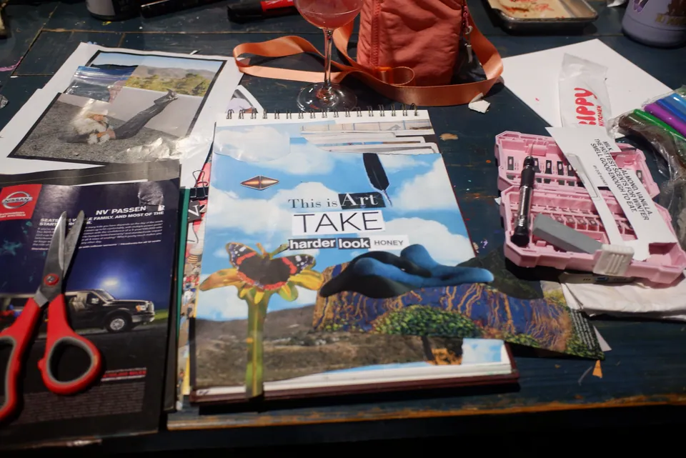 Her table at night: a sketchbook holding a collage with the words This is Art, TAKE a harder look, among red scissors, a pink paint palette and open magazines.