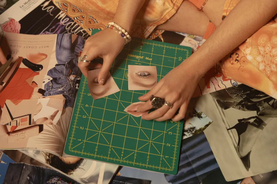 From above, Camila's hands place a cut-out eye and a pair of lips on a mostly empty green cutting mat, orange patterned clothing behind.