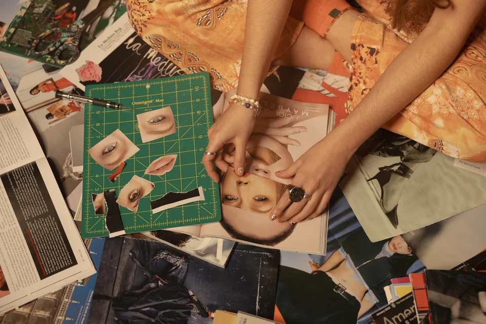 Camila sits cross-legged among magazines, lifting a cut piece from a model's face on an open page, cutting mat of eyes and lips beside her.