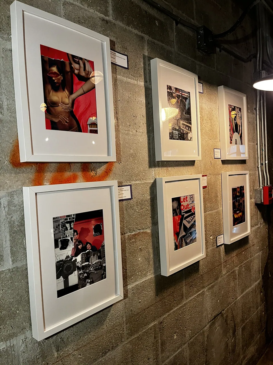Six collages in white frames on a block wall marked with orange paint, each with a small label beside it.