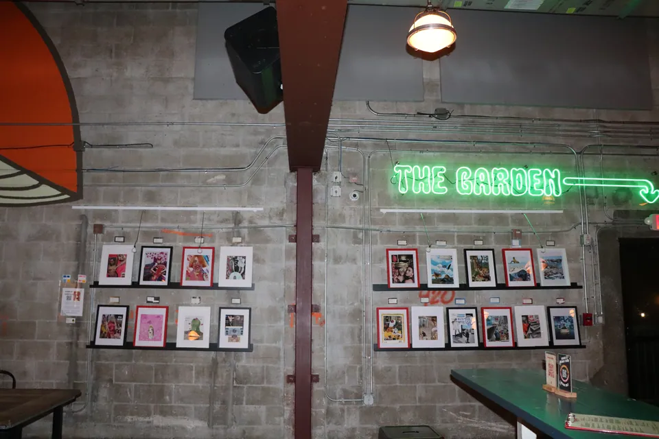 A block wall at the brewery hung with two groups of framed collages on ledges, under a green neon sign.